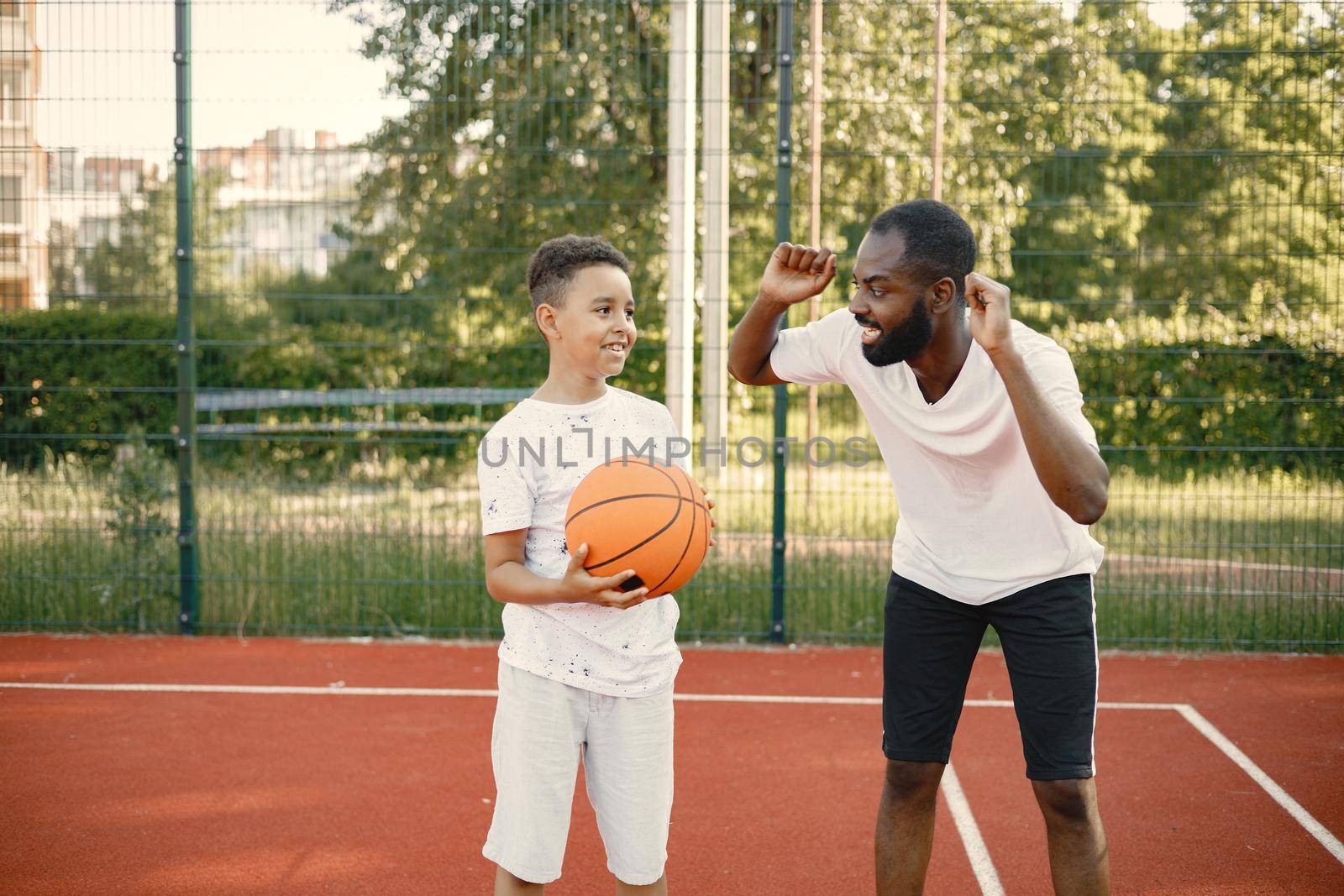 Young father and his son playing basketball in basketball court near the park. Man and boy wearing white t-shirts. Man has his arms up and cheering.