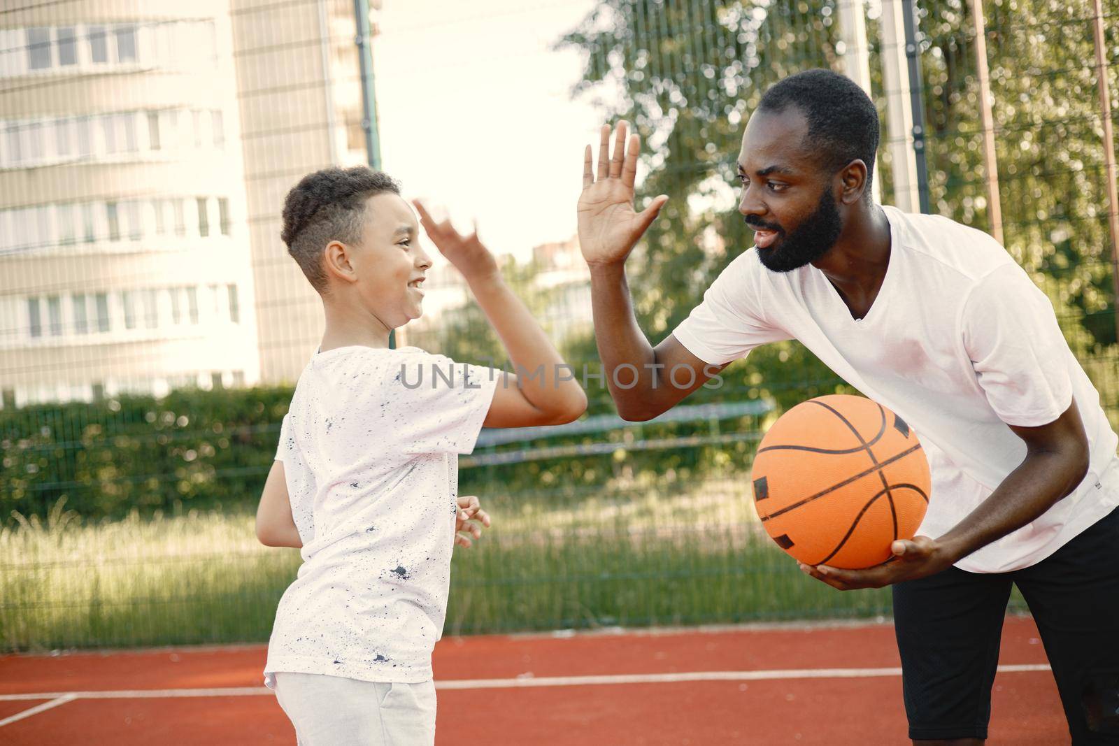 Young father and his son playing basketball in basketball court near the park. Man and boy wearing white t-shirts. They give a high five to each other.