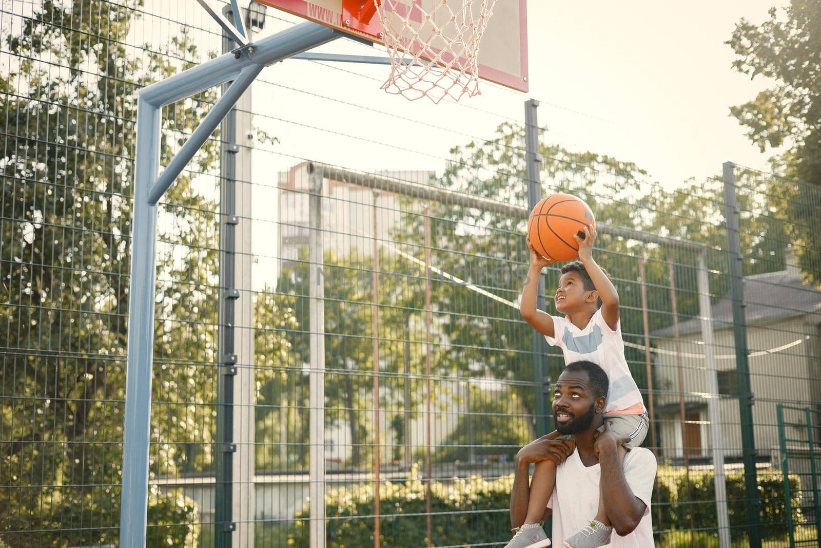 Black father holding on shoulders his little son to help him to score a basket. They standing on a basketball court. Man and boy wearing white t-shirts.