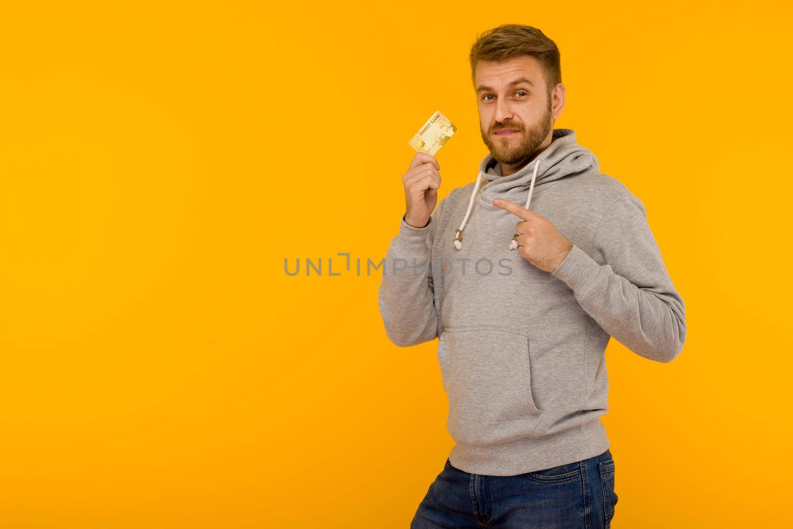 Attractive man in a gray hoodie points a finger at the credit card that is holding in his hand on a yellow background by zartarn