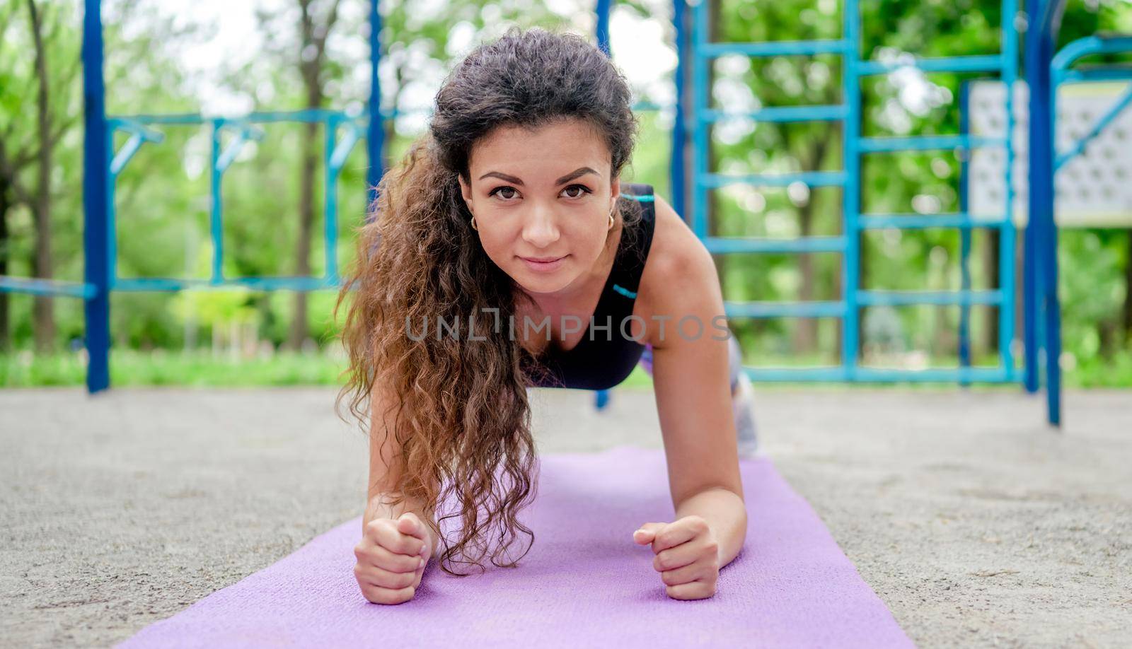 Sport girl doing plank exercise on yoga mat outdoors and looking at camera. Young woman during workout at stadium