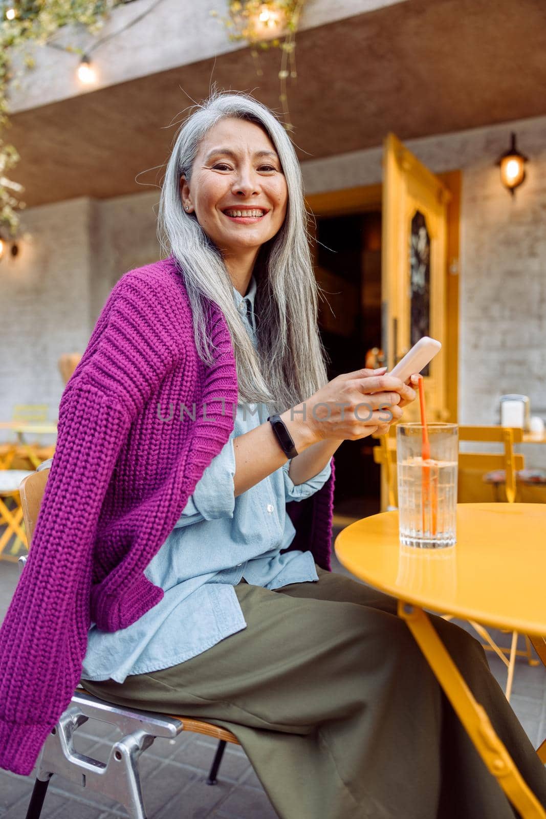 Happy senior Asian woman in purple jacket holds mobile phone sitting at small yellow table on outdoors cafe terrace on autumn day