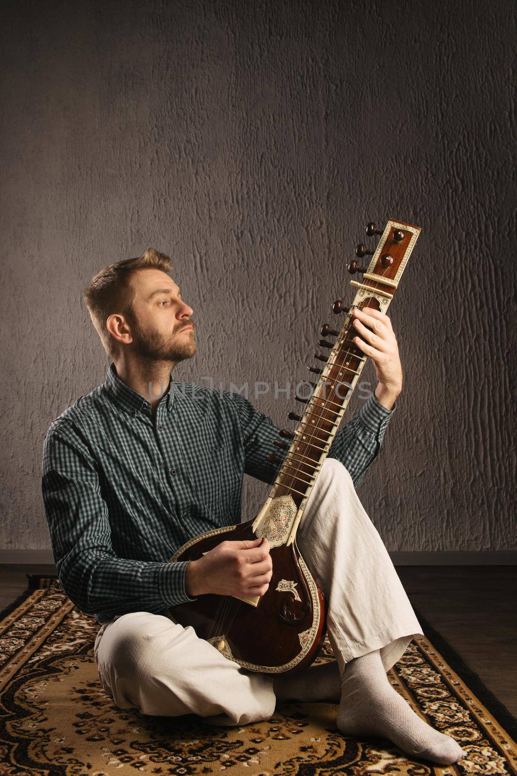 Portrait of a European man playing the sitar sitting on the carpet- Image