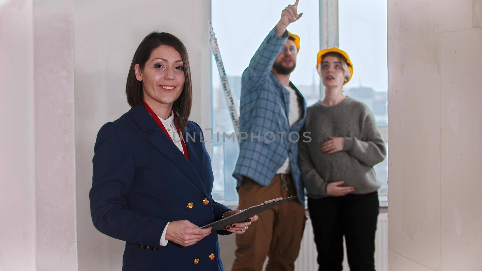 A smiling real estate agent standing in draft apartment - young married couple looking around on the background. Mid shot