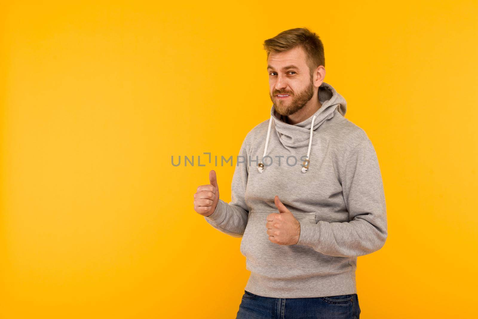 Handsome attractive European man in gray hoodie thumb up on yellow background - image