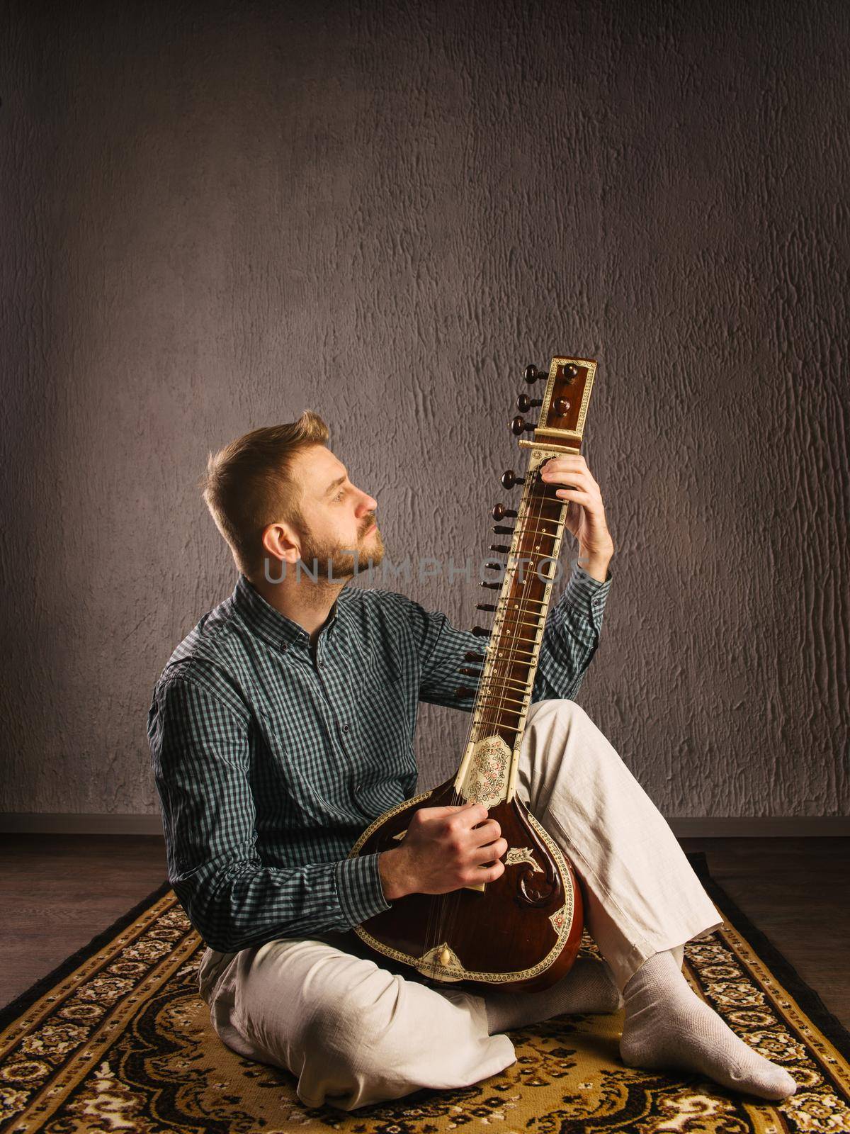 Portrait of a European man playing the sitar sitting on the carpet- Image