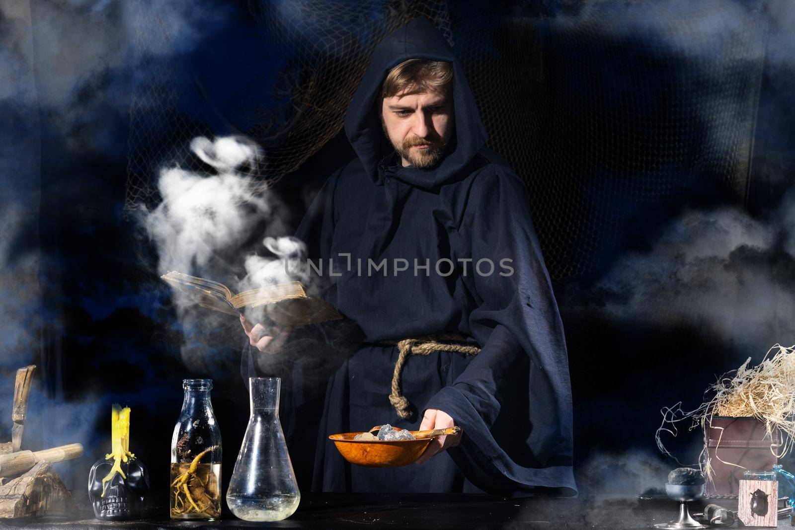 The medieval alchemist make magic ritual at the table in his smoke laboratory. Halloween concepy background.