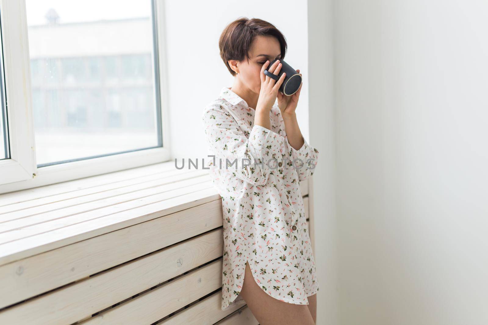 Beautiful young woman with cup of tea standing near the window at home