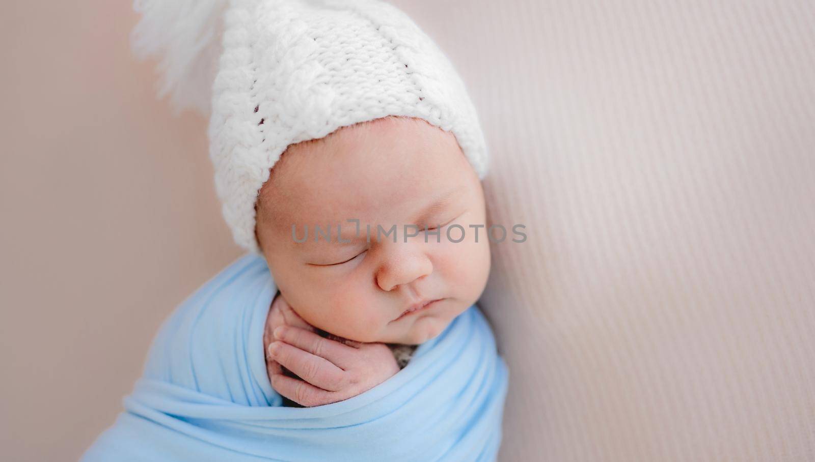 Newborn baby boy sleeping wearing hat and holding tiny hands under cheeks. Infant child kid resting during studio photoshoot