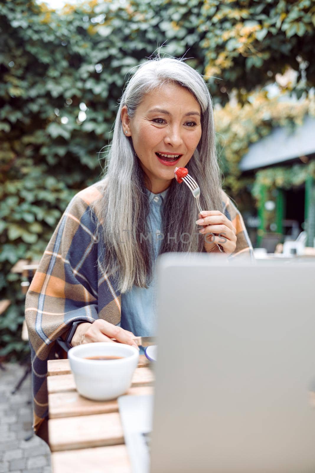 Beautiful senior Asian woman eats fresh strawberry with cream in front of open laptop at table on outdoors cafe terrace on autumn day