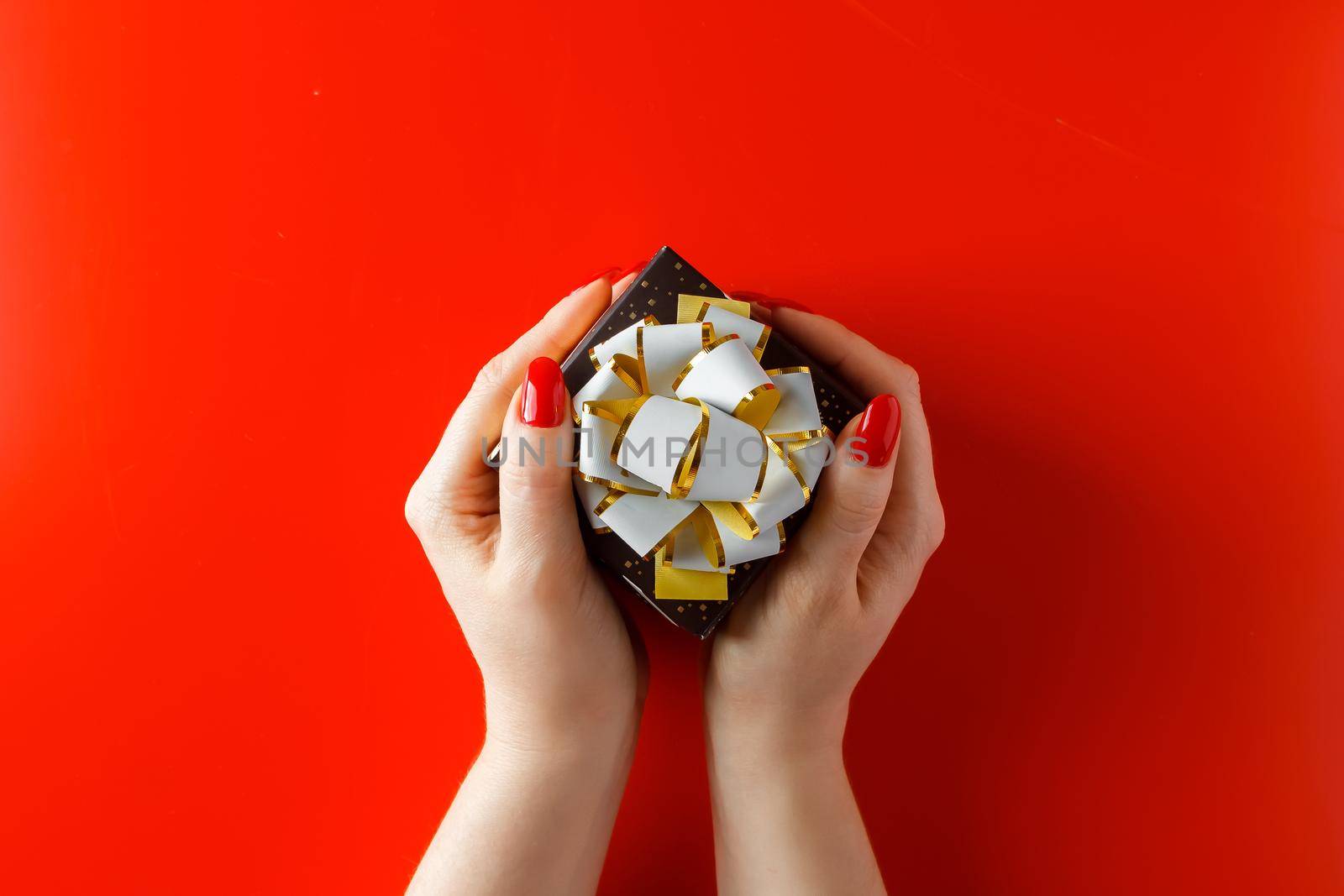 Gift with a red ribbon in hands on a white background. Beautiful little gift with a ribbon in the hands of a woman on a red background. Giving a present for the holiday.