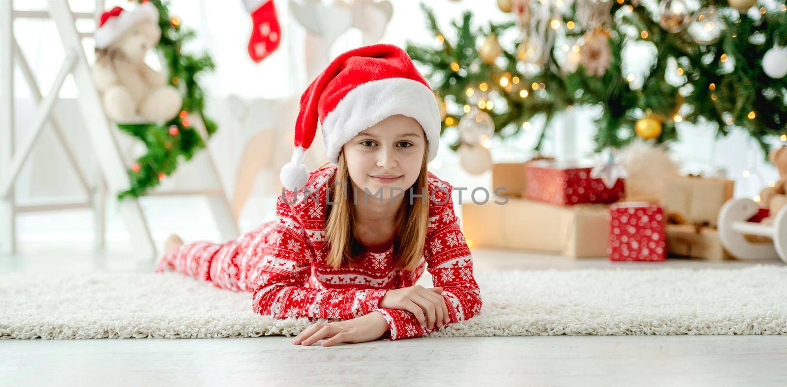 Pretty child girl lying on floor at Christmas home with traditional decorated tree. Happy smiling kid in New Year time