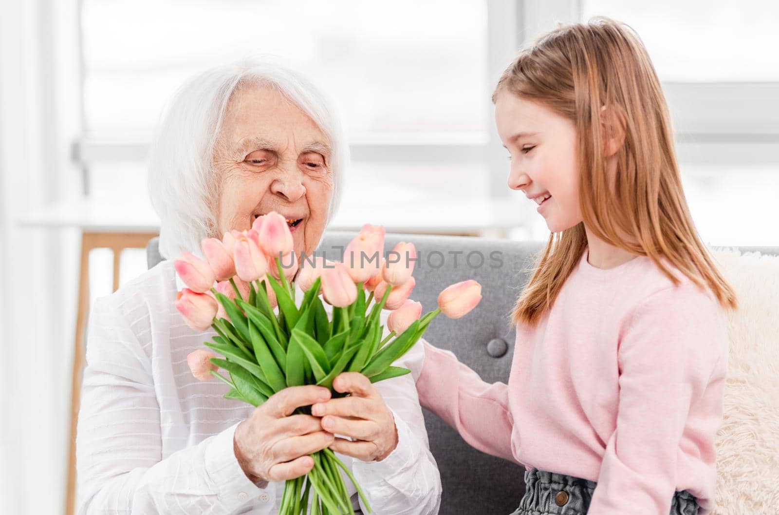 Grandmother sitiing on the sofa with preschool granddaughter, looking on flowers tulips bouquet and smiling