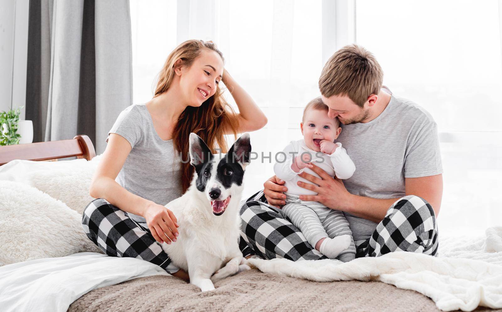 Beautiful family with baby boy sitting on the bed with cute dog and smiling. Mother and father with their son and doggy togetherin the morning. Beautiful parenthood time. Pet with owners