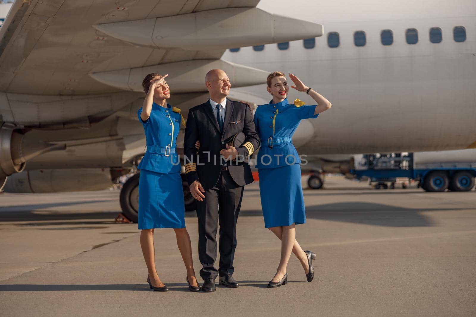 Full length shot of happy pilot standing together with two stewardesses in bright blue uniform in front of an airplane on a sunny day by Yaroslav_astakhov