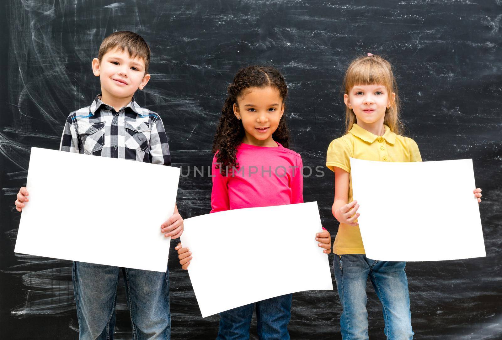 three cute children with empty paper sheets in hands and chalky blackboard on background
