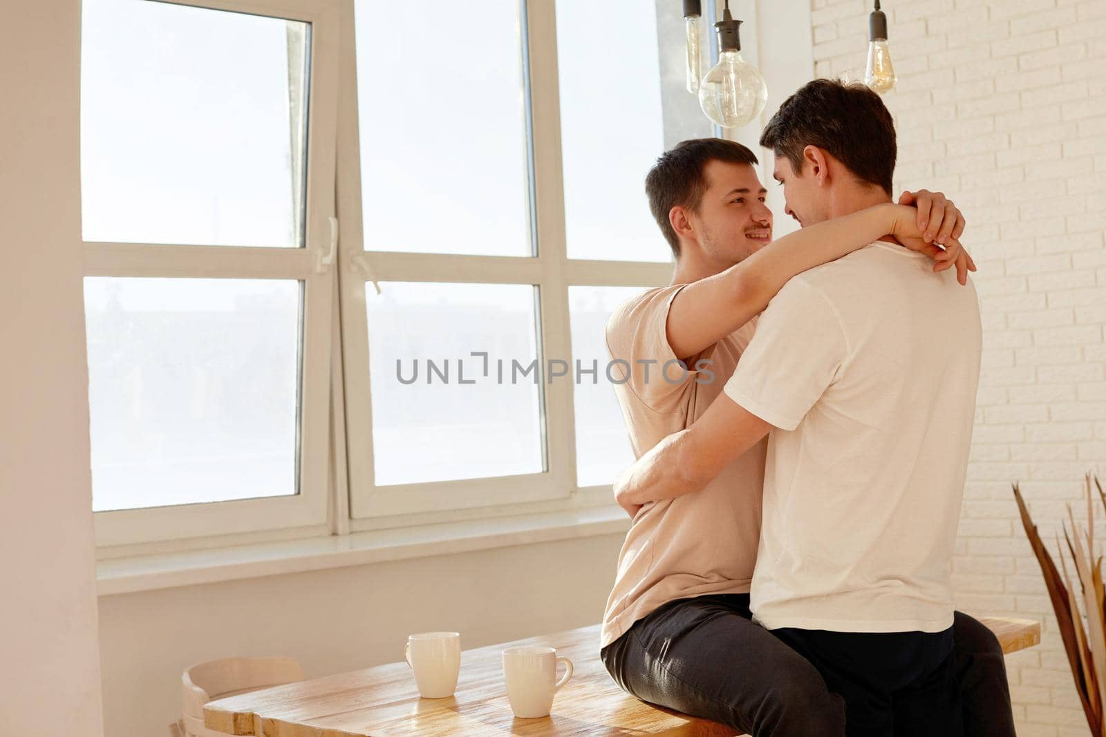 Cute male gay couple hugging at home in the morning while having breakfast while sitting on the table against the background of a white dining room