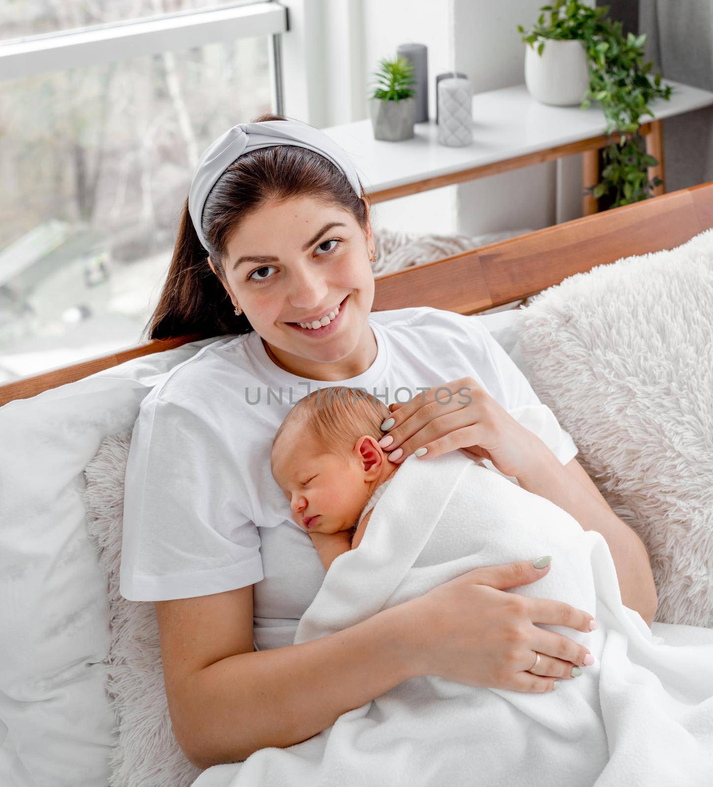 Young mother with napping newborn baby staying in the bed and smiling. Portrait of beautiful girl with her child. Mom holding sleeping infant child on her chest