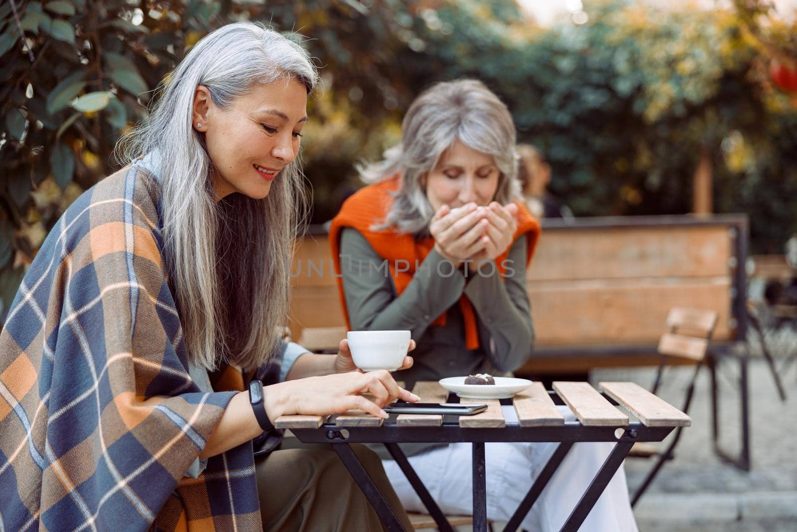 Senior Asian woman uses mobile phone while hoary haired friend drinks tea sitting at small wooden table in street cafe on nice autumn day