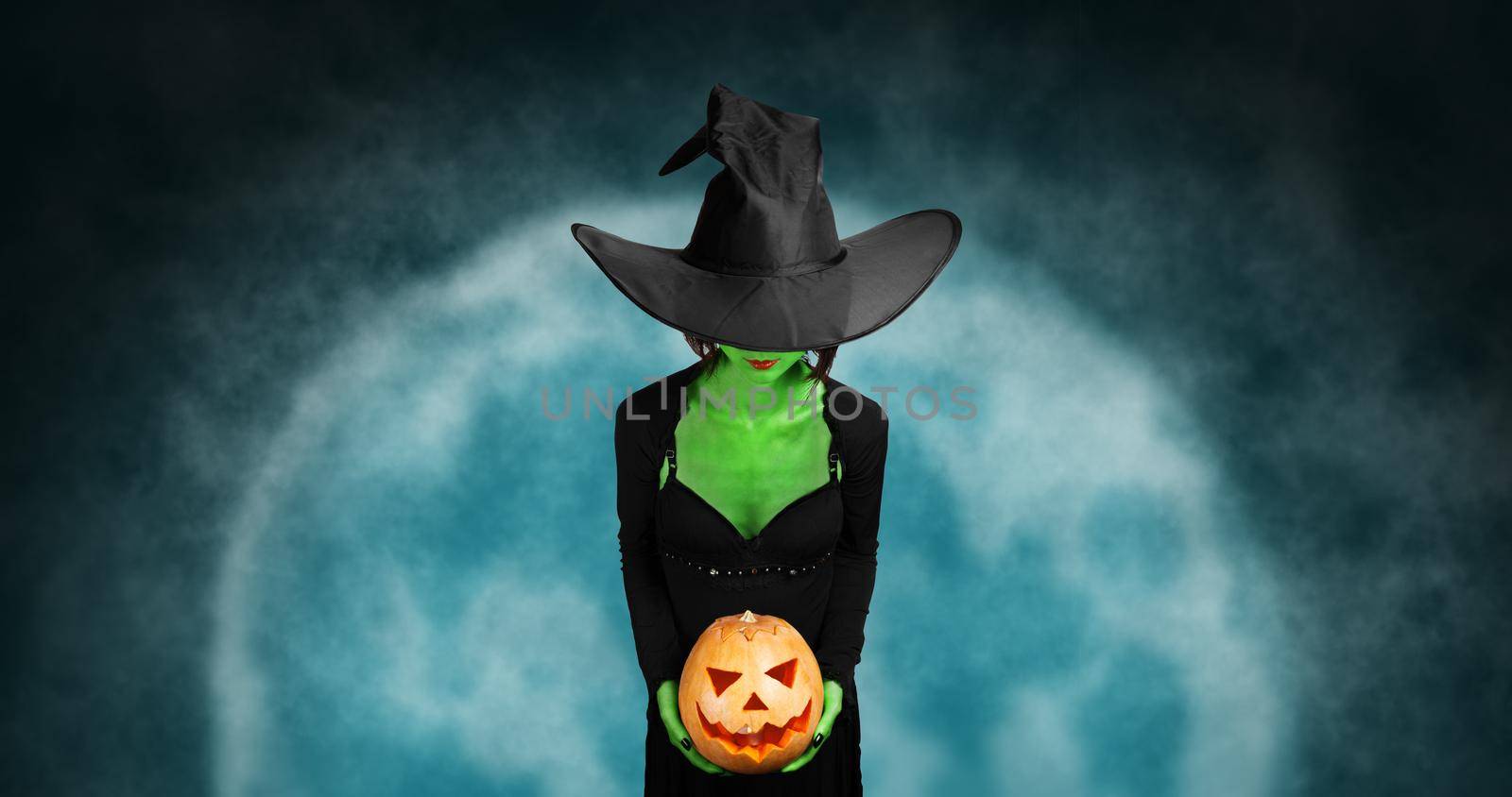 Green witch holds Halloween carved pumpkin on background of full moon. Halloween, horror theme