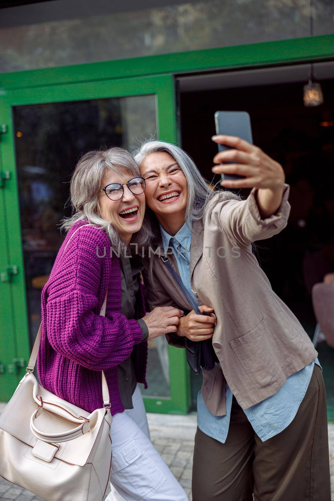Beautiful laughing mature Asian lady with companion take selfie with smartphone on modern city street. Long-time friendship relationship