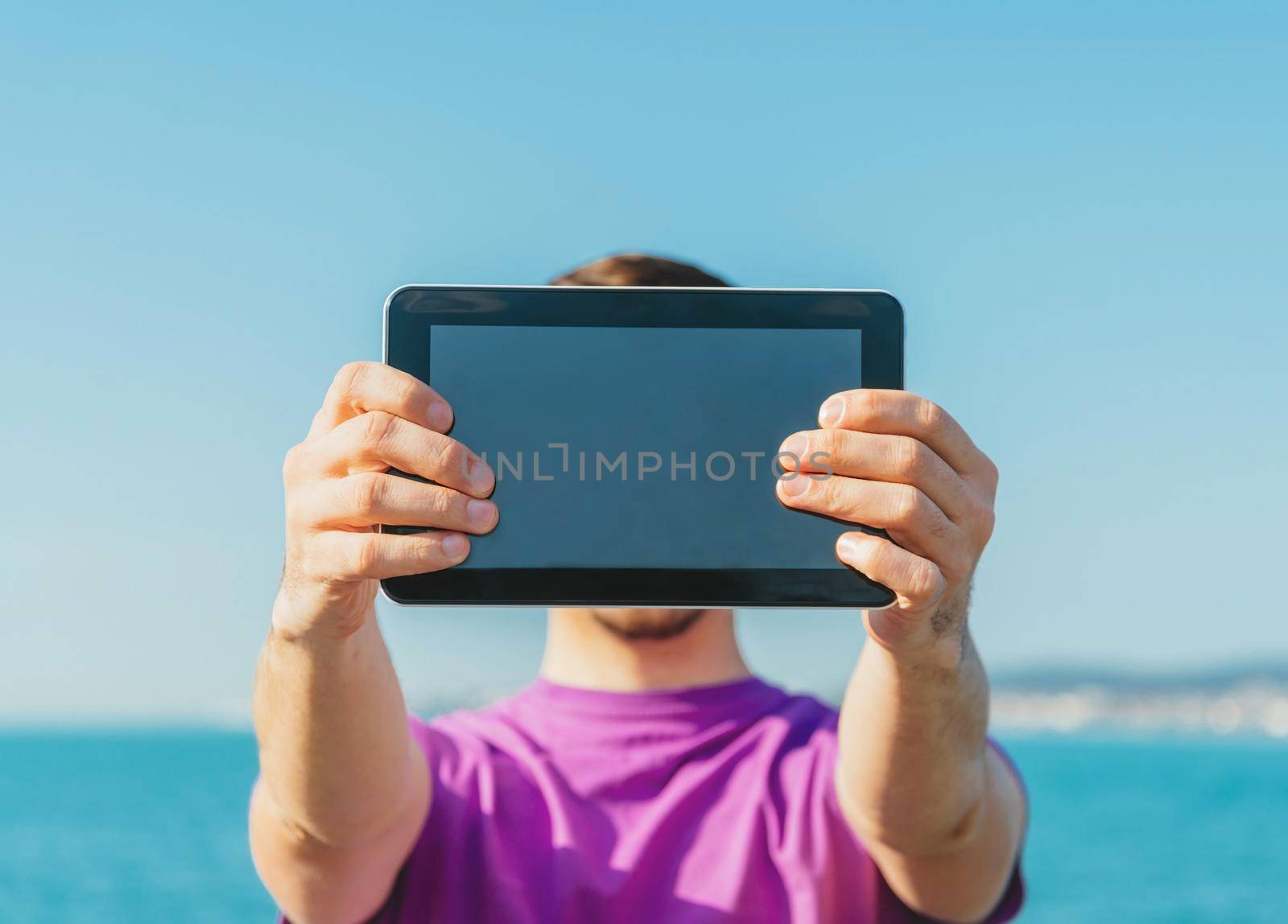 Young man holding digital tablet in front of him on beach in summer, space for text on screen of tablet