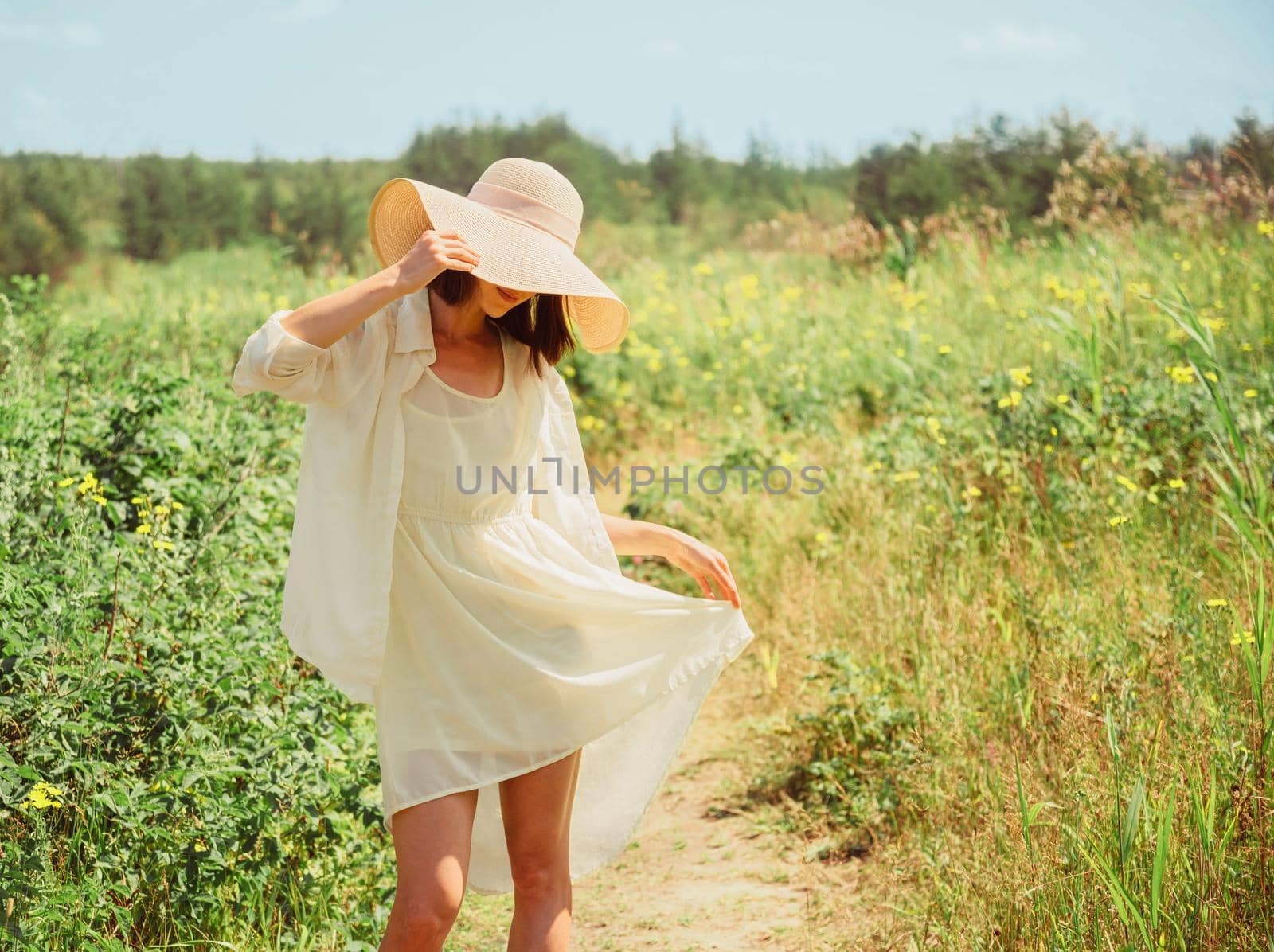 Happy beautiful young woman in yellow dress and hat with wide brim walking in summer park