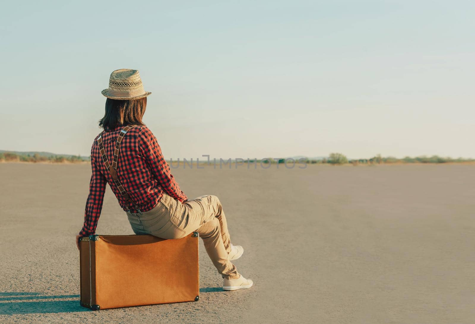 Traveler young woman in a hat sitting on suitcase on road. Space for text in left part of image