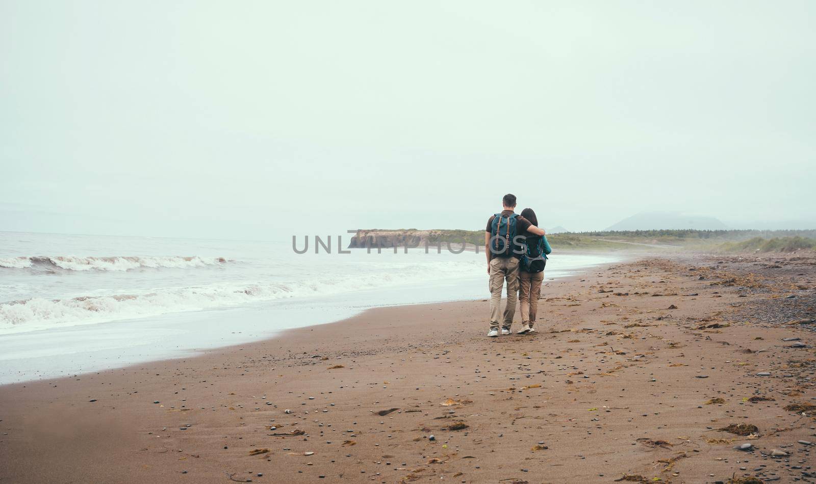 Traveler loving couple with backpacks walking on beach near the sea in summer. Man embracing a woman, rear view