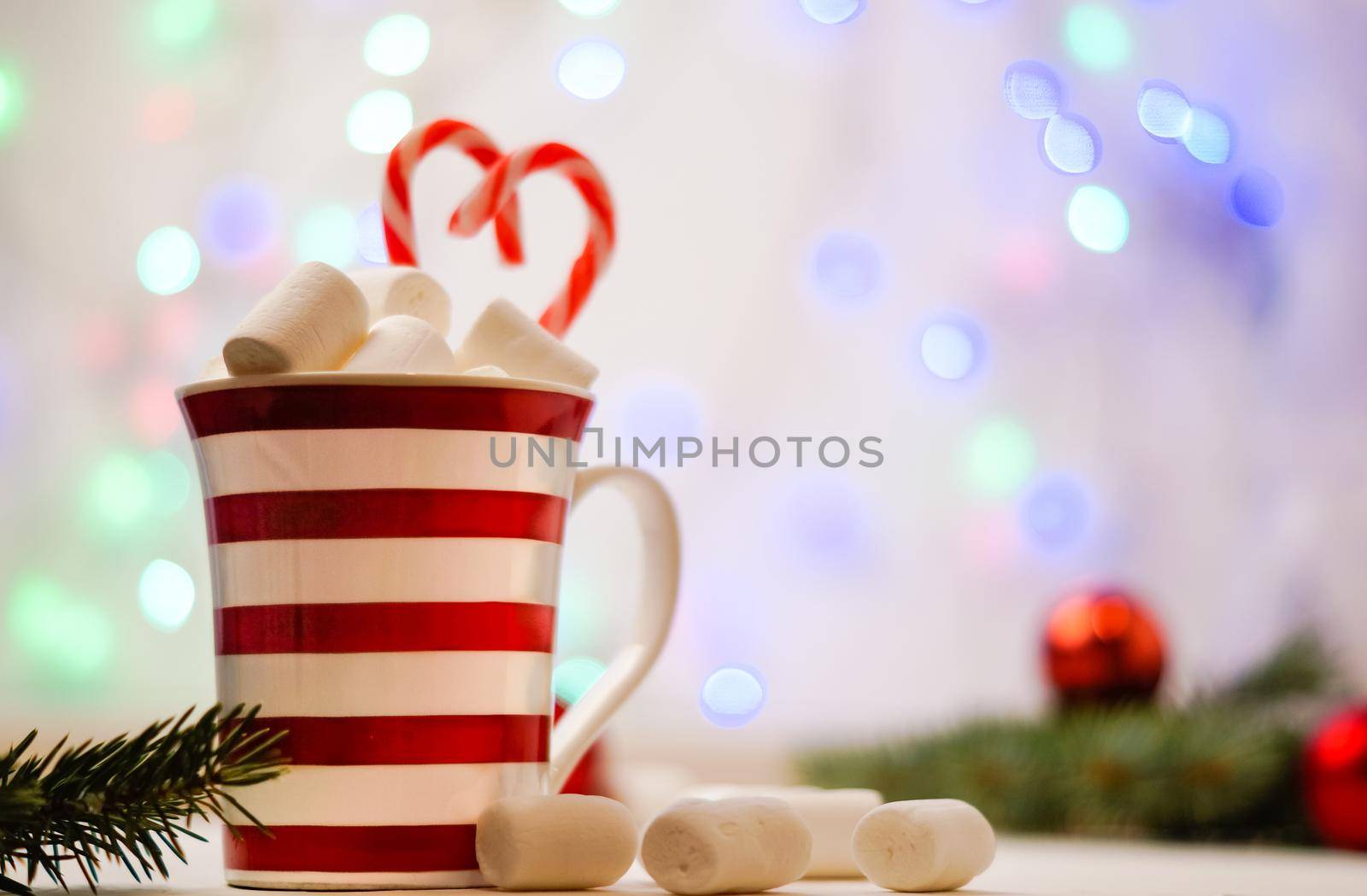 Hot cocoa with marshmallows and red-white lollipop. Hot chocolate with marshmallows in a New Year's cup against a background of multi-colored bokeh