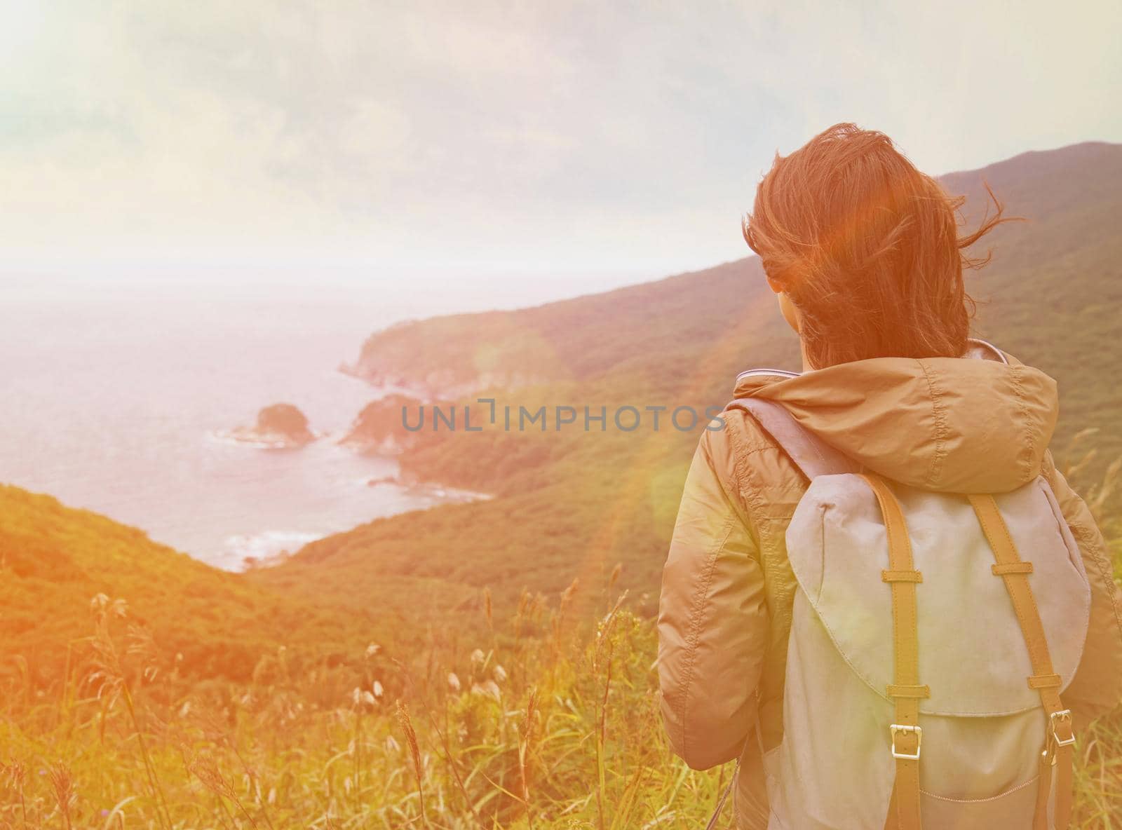 Hiker young woman with backpack looking into distance to the island in summer. Image with sunlight effect