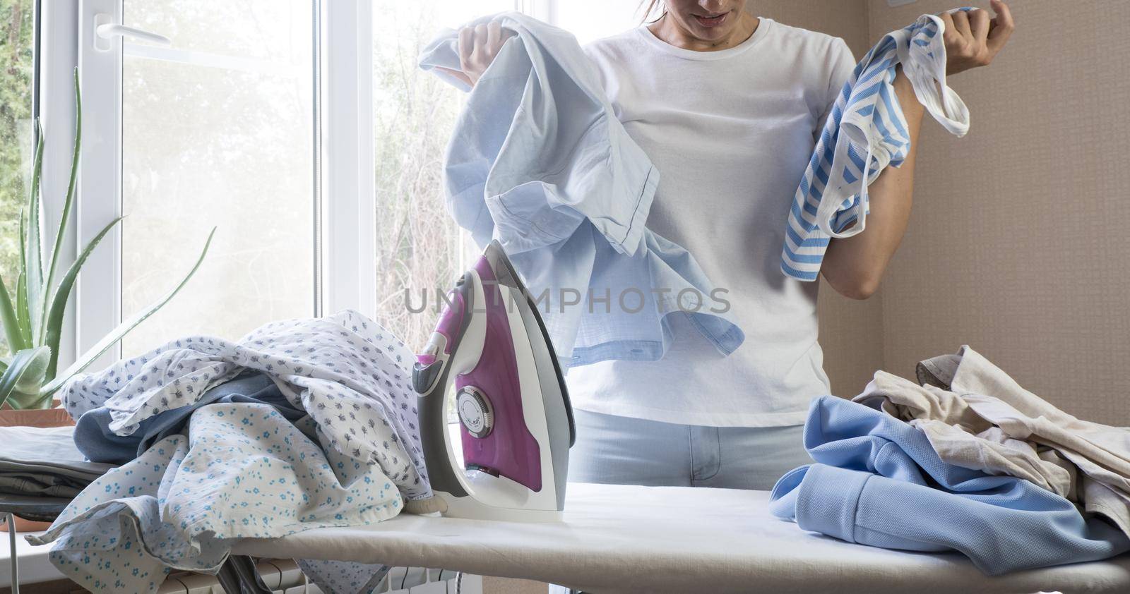 Frustrated tired young woman holds stares at iron and pile of ironed clothes at home. Housework.