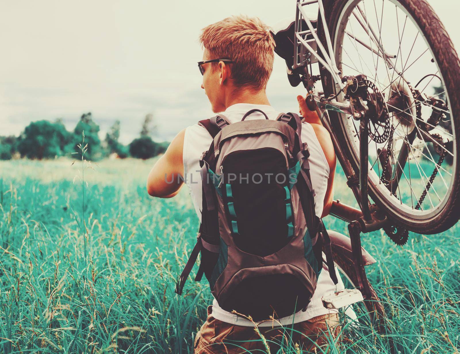 Cyclist man with backpack carries bicycle in green meadow in summer. Image with instagram filter