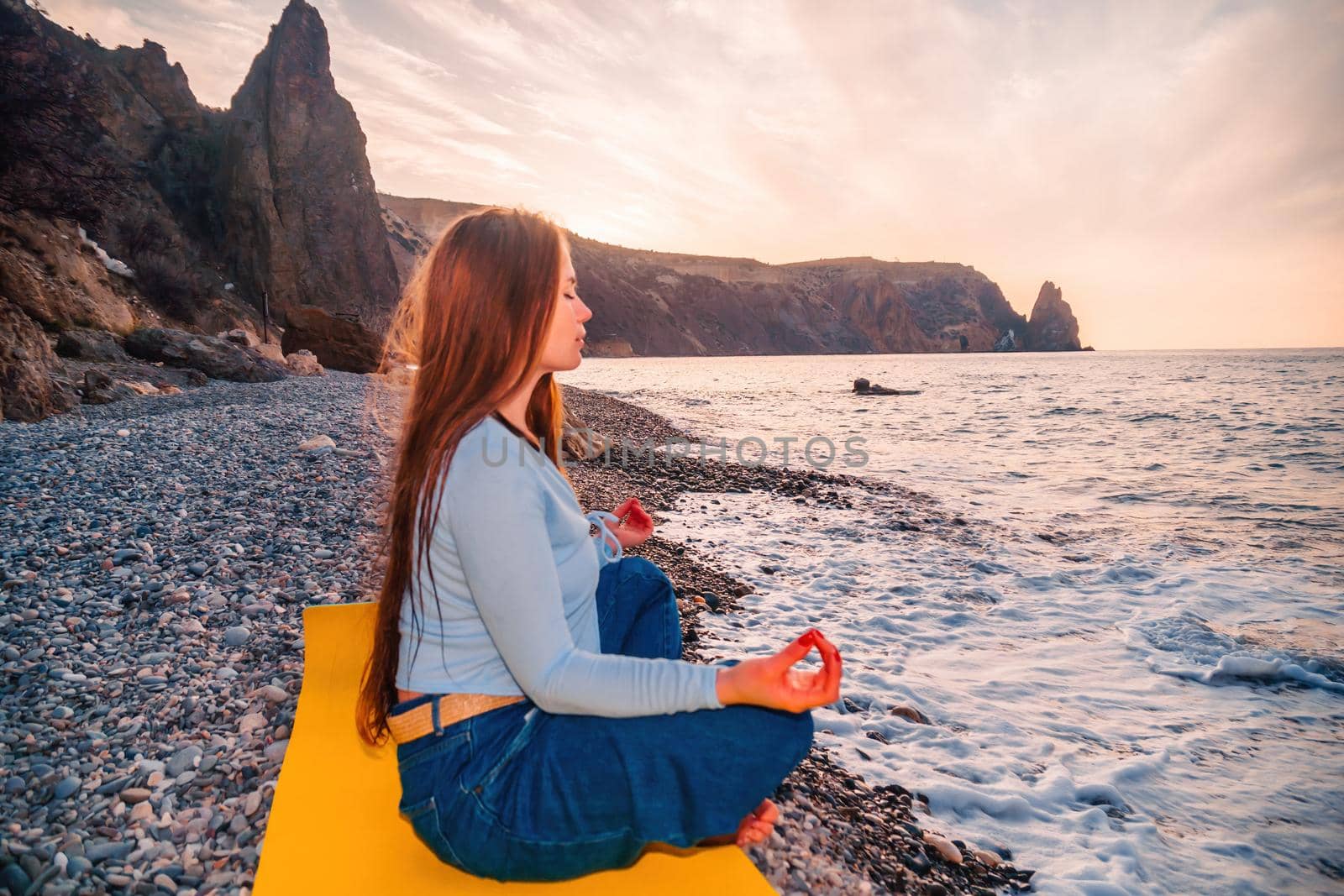 Young woman in swimsuit with long hair practicing stretching outdoors on yoga mat by the sea on a sunny day. Women's yoga fitness pilates routine. Healthy lifestyle, harmony and meditation concept.