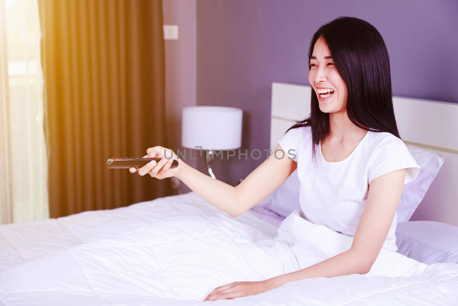 woman watching TV laughing having fun with remote control on the bed