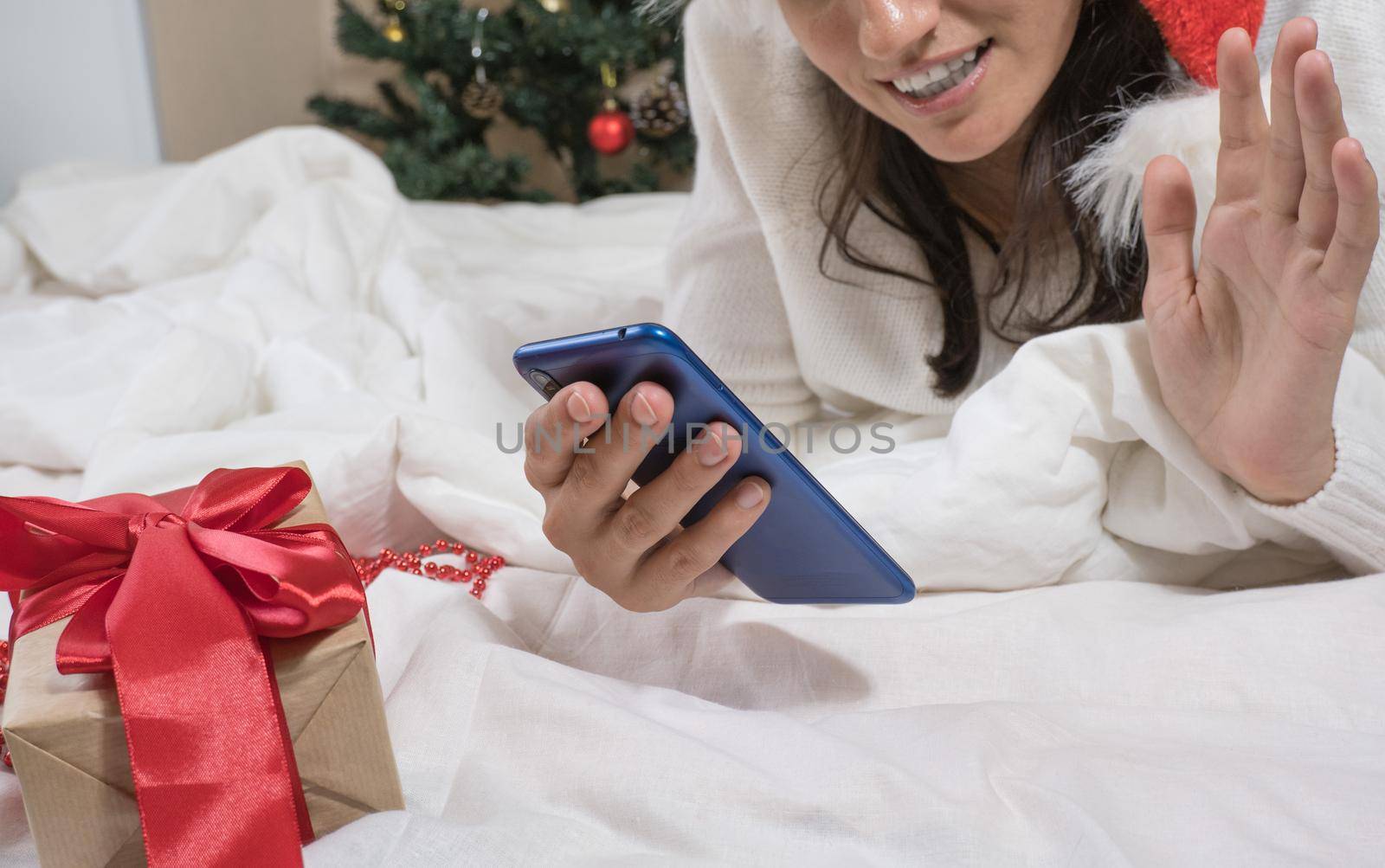 Beautiful happy brunette woman makes a selfie by phone in a white sweater and a Christmas hat and a Christmas present while lying in a white bed.