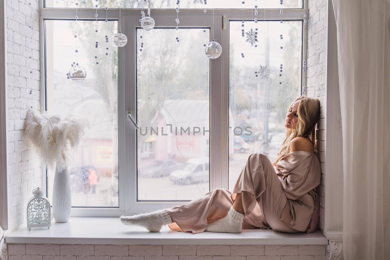 Resting and thinking woman in pajamas sitting on the window-sill at home. Side view