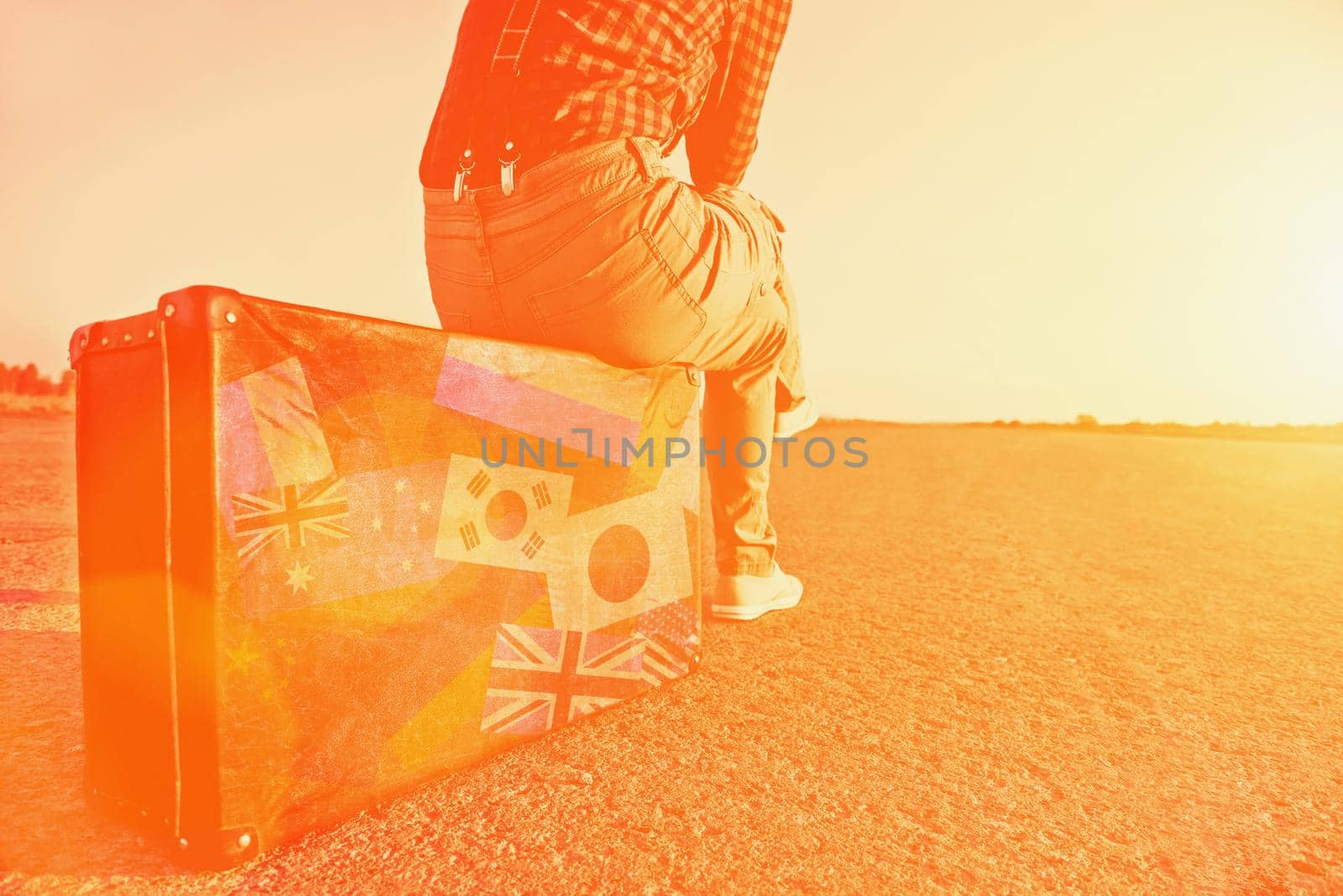 Tourist woman sitting on a suitcase on road. Suitcase with stamps flags of different countries. . Image with sunlight effect
