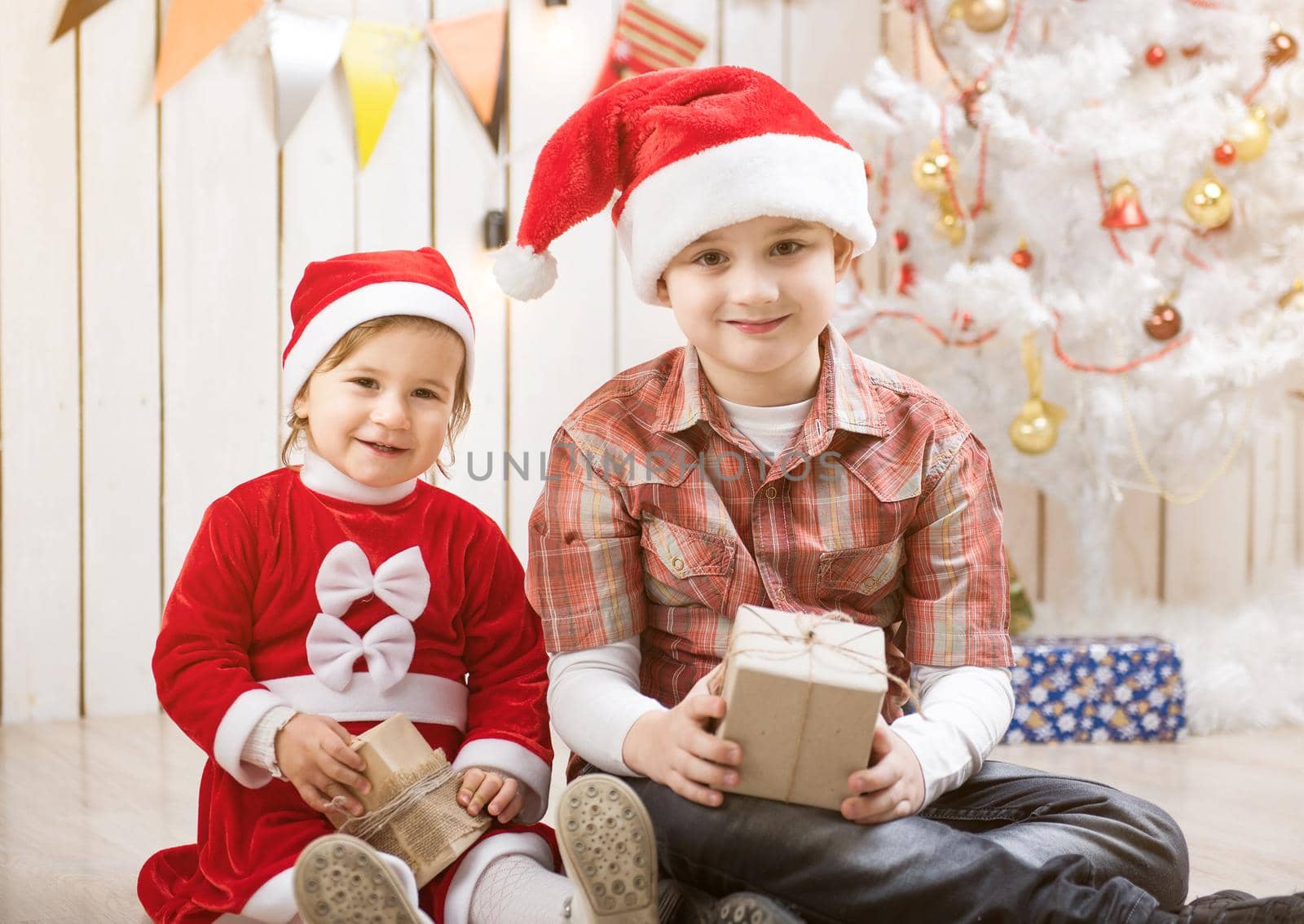 Kids in red santa hats sitting in decorated room with present boxes in hands
