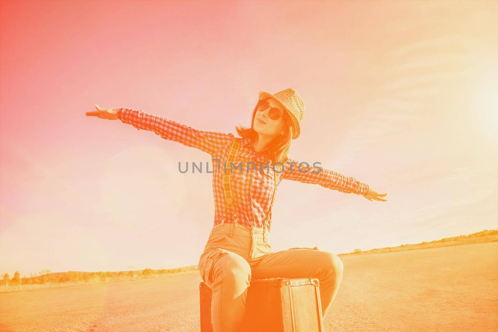 Happy hipster girl sits on vintage suitcase on road and makes a gesture of flight. Image with sunlight effect