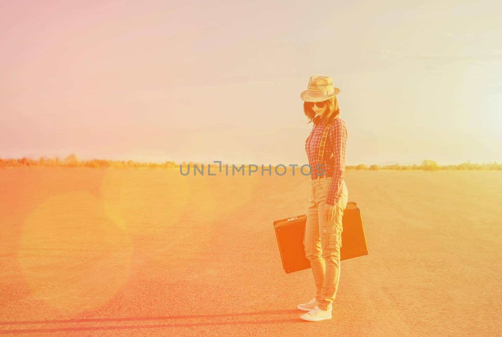 Traveler young woman in hat and sunglasses standing with a suitcase on road in summer. Image with sunlight effect