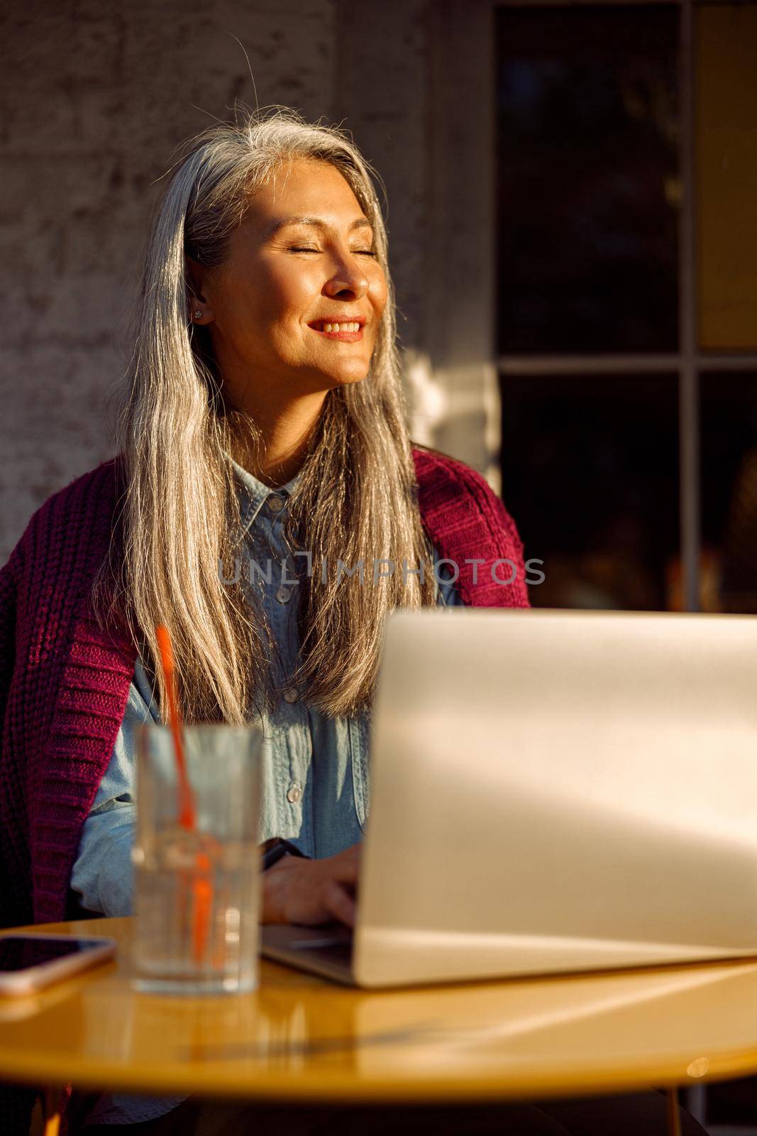 Cheerful senior Asian lady with works on contemporary laptop witting at small round table on outdoors cafe terrace at sunset