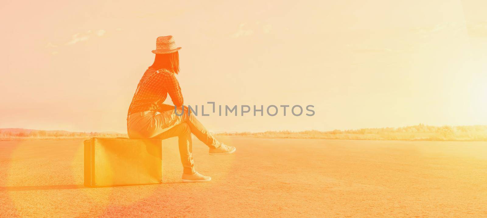 Traveler woman sits on a suitcase and looks away on road. Image with sunlight effect