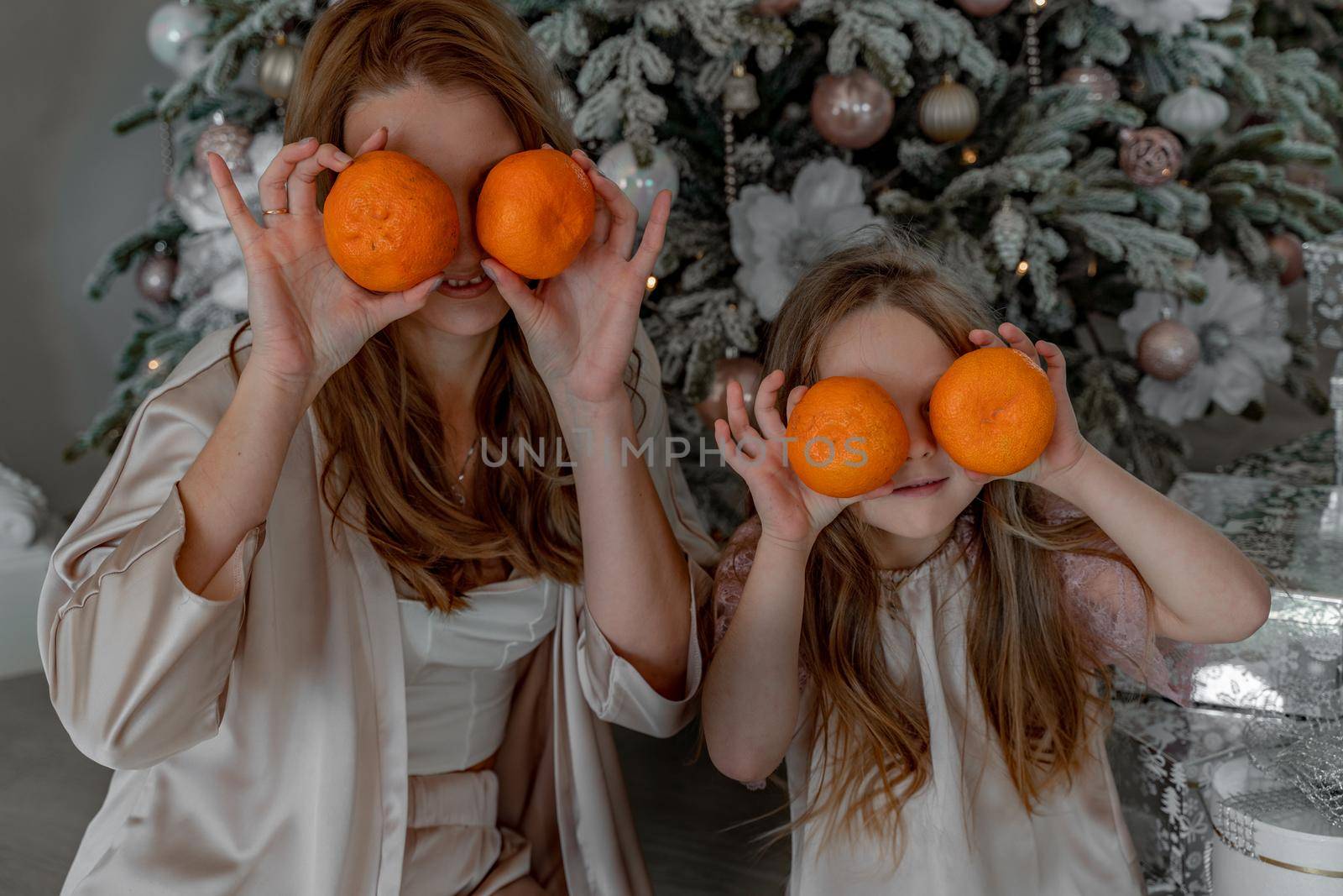 Mom and daughter hold tangerines at eye level instead of glasses. They are sitting under a decorated Christmas tree in their pajamas. The concept of Christmas, family holidays by Matiunina