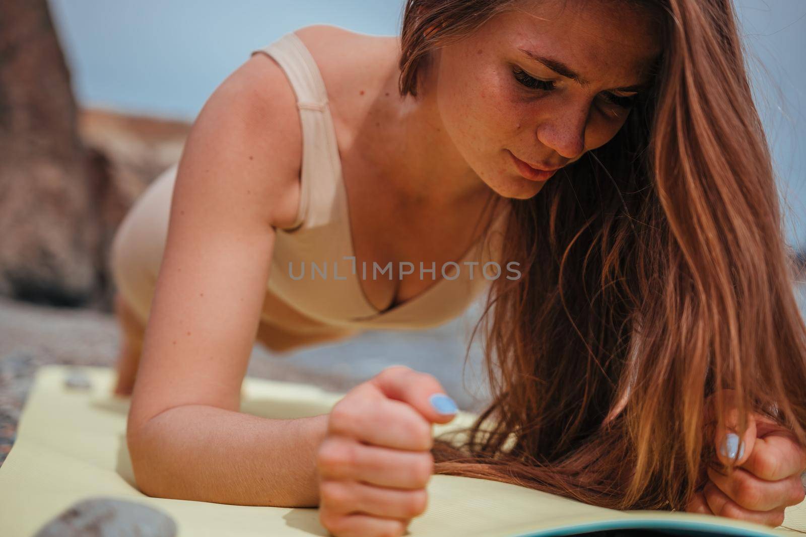 Young woman in swimsuit with long hair practicing stretching outdoors on yoga mat by the sea on a sunny day. Women's yoga fitness pilates routine. Healthy lifestyle, harmony and meditation concept.