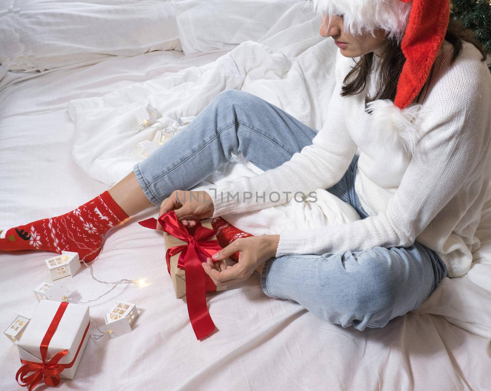 Happy young brunette woman in santa claus hat opens a gift box for Christmas lying on a white bed. Christmas gifts