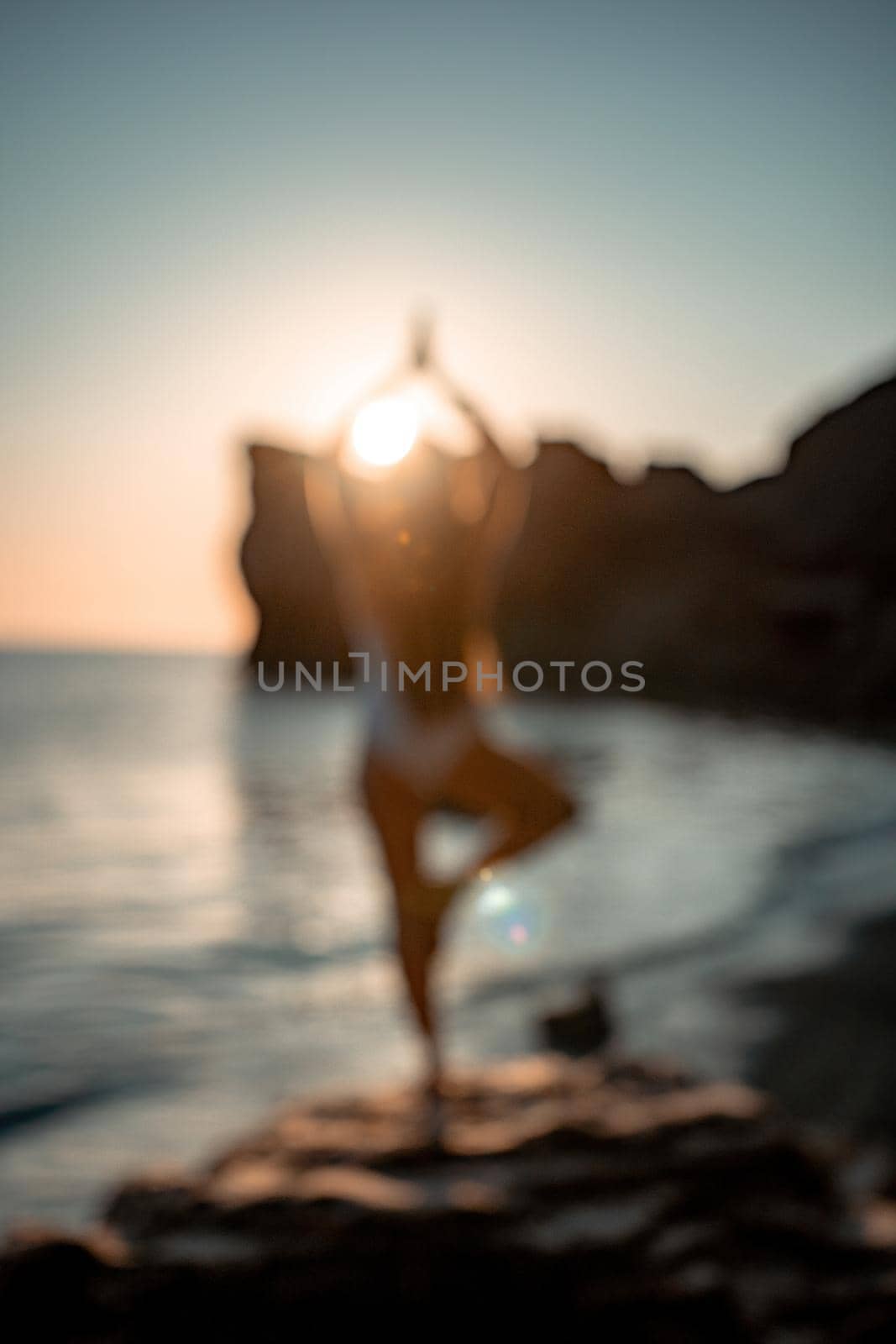 Defocused silhoute of Young woman with long hair in white swimsuit practicing outdoors on yoga mat by the sea on a sunset. Women's yoga fitness routine. Healthy lifestyle, harmony and meditation by panophotograph