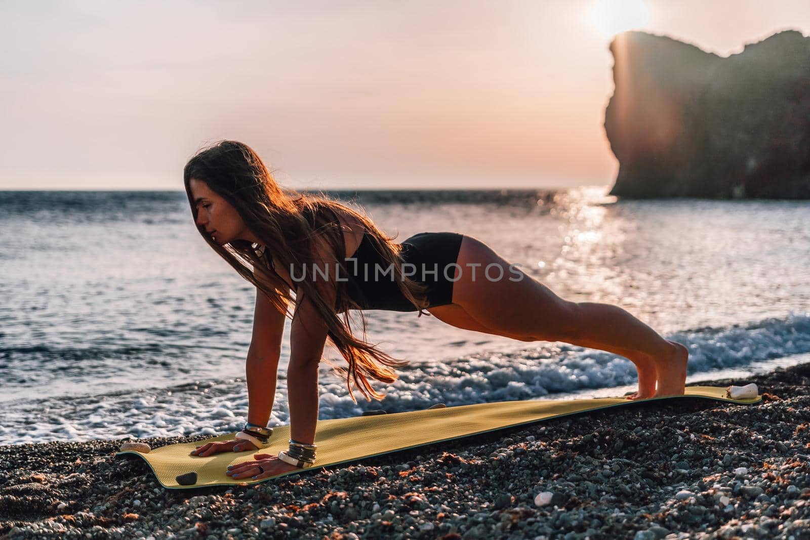 Young woman in swimsuit with long hair practicing stretching outdoors on yoga mat by the sea on a sunny day. Women's yoga fitness pilates routine. Healthy lifestyle, harmony and meditation concept.