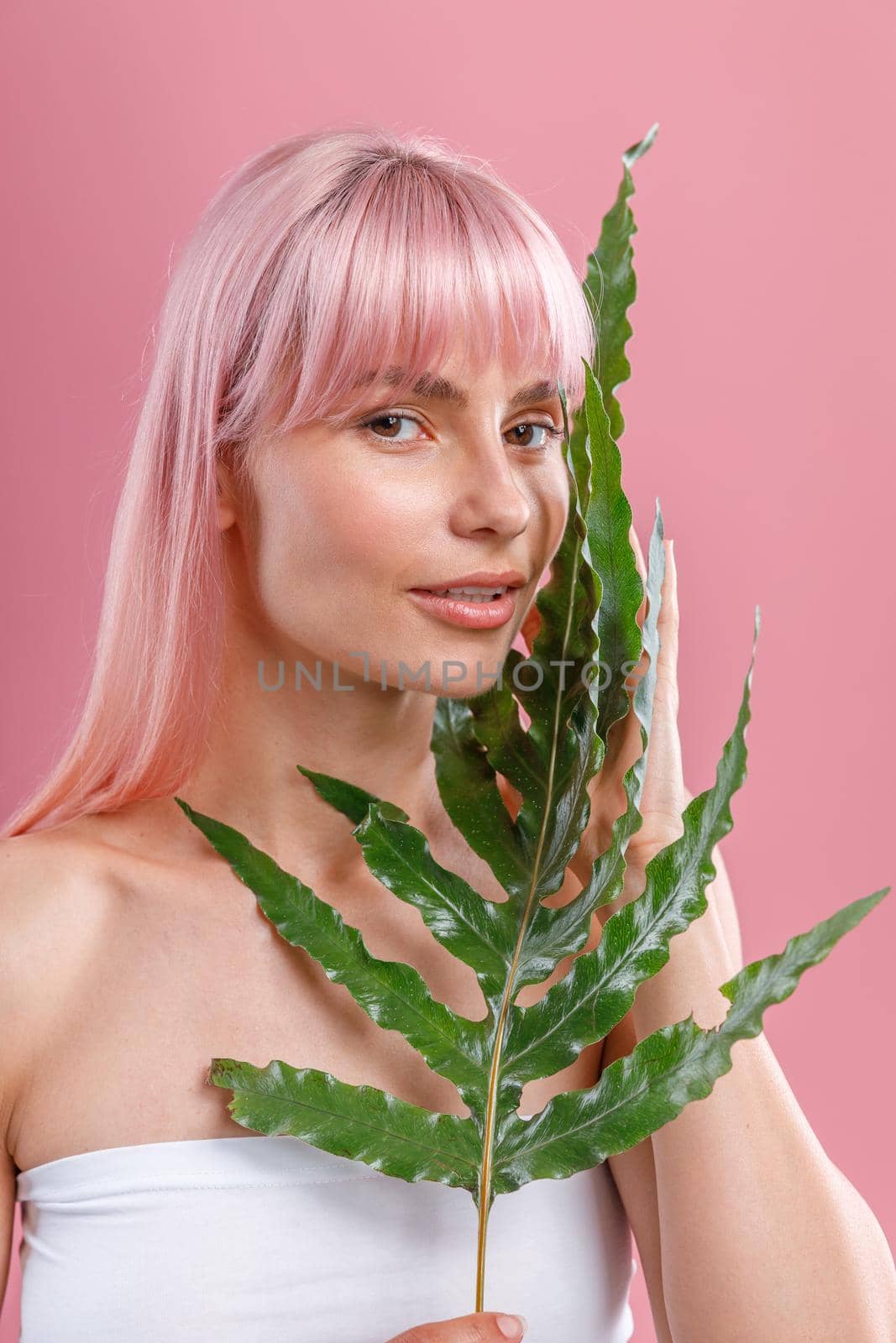 Portrait of beautiful woman with pink hair and perfect skin looking at camera, posing with plant leaf isolated over pink studio background. Beauty, skin care, natural cosmetics concept