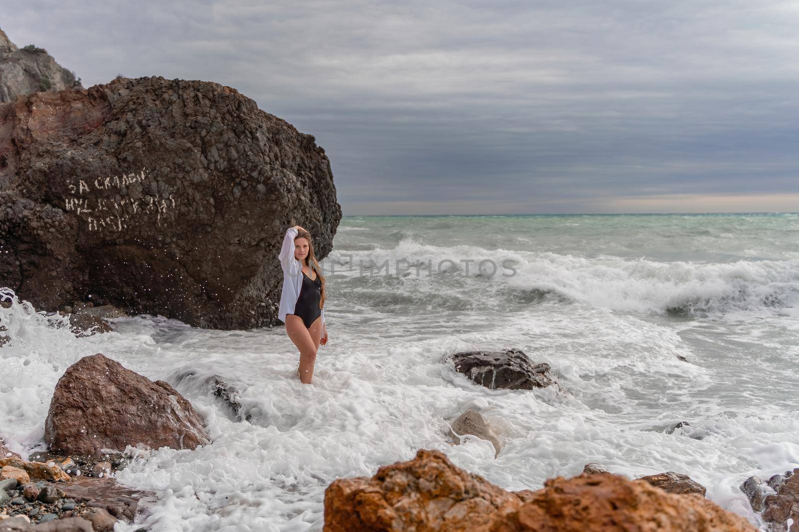 A beautiful girl in a black dress is walking on the waves, big waves with white foam. A cloudy stormy day at sea, with clouds and big waves hitting the rocks